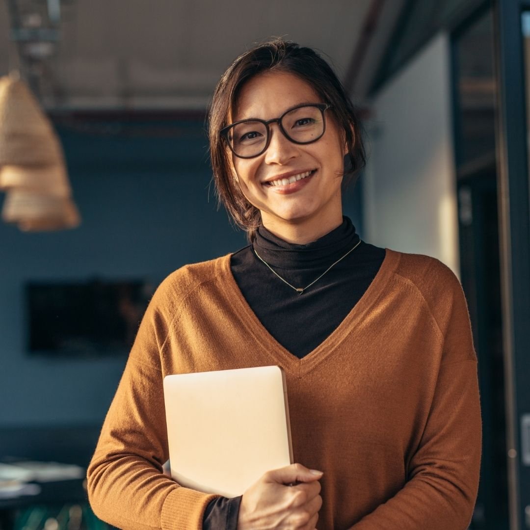 Woman in turtleneck and sweater smiling in office while holding laptop